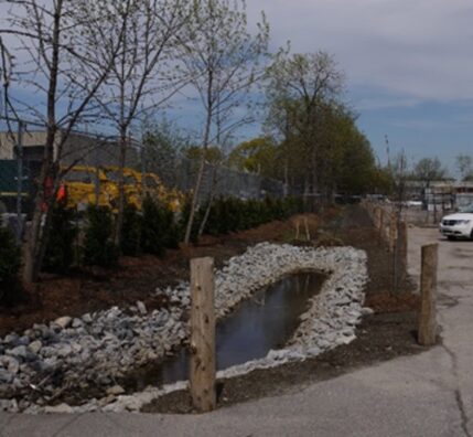 A swale in a commercial parking lot at the property boundary filled with rock and ponding water.