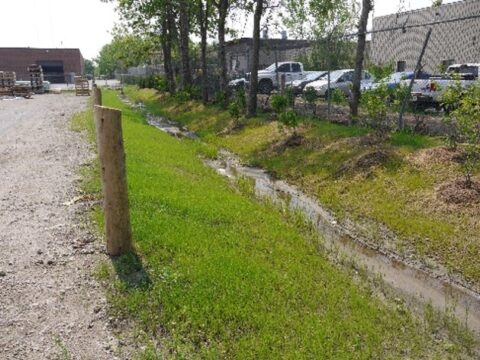A swale at the edge of a property situated between a gravel driveway and chain link fence.
